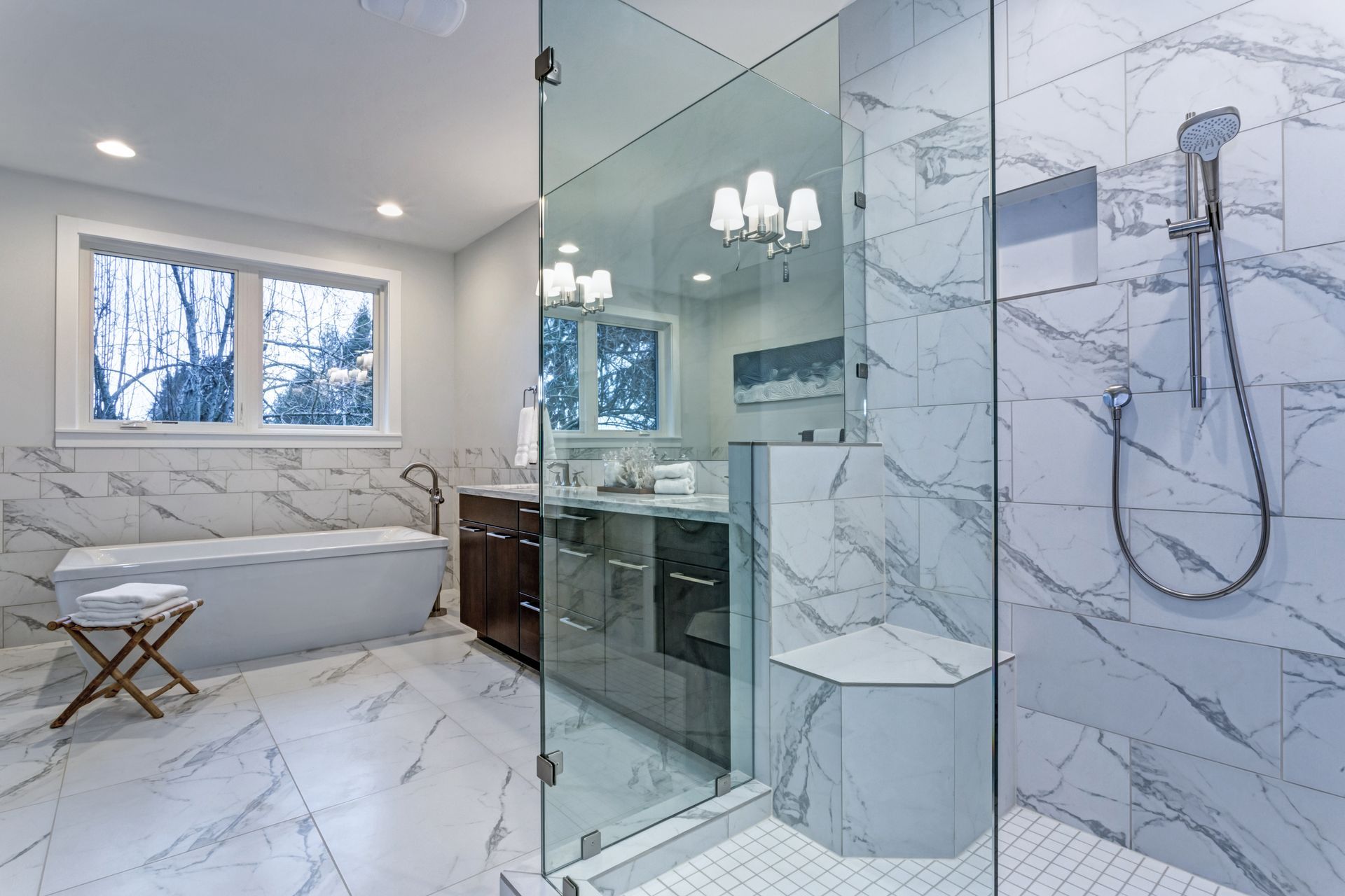 Modern bathroom with marble-look tile, glass shower, soaking tub, dark wood vanity, and a window with winter view.
