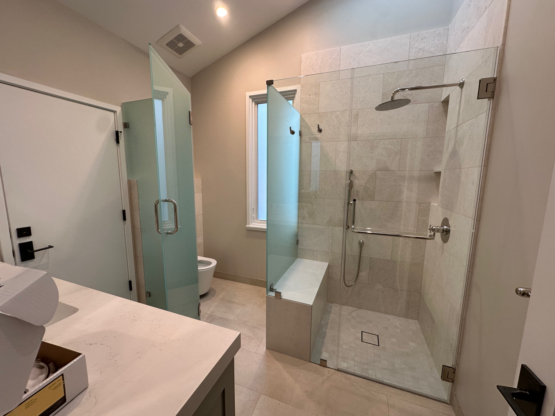 Bathroom with glass shower, bench, and toilet. Beige tile and neutral walls.