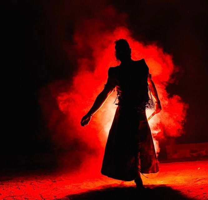 Sagoma di una persona con indosso un lungo abito, braccia tese, illuminata da luce rossa e fumo in uno spazio buio.