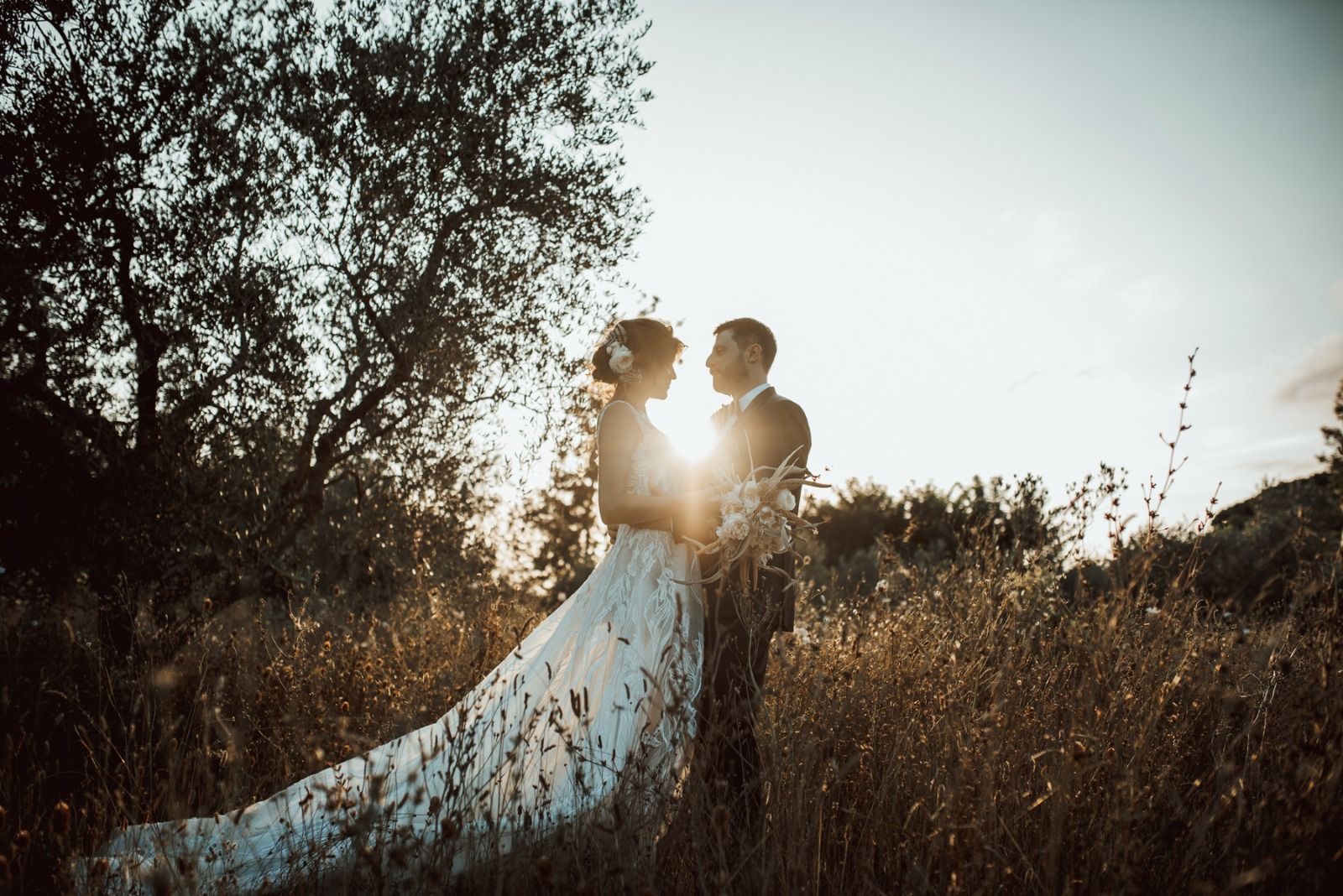 Una coppia si abbraccia in un campo al tramonto. La sposa indossa un abito bianco svolazzante, lo sposo un abito elegante.