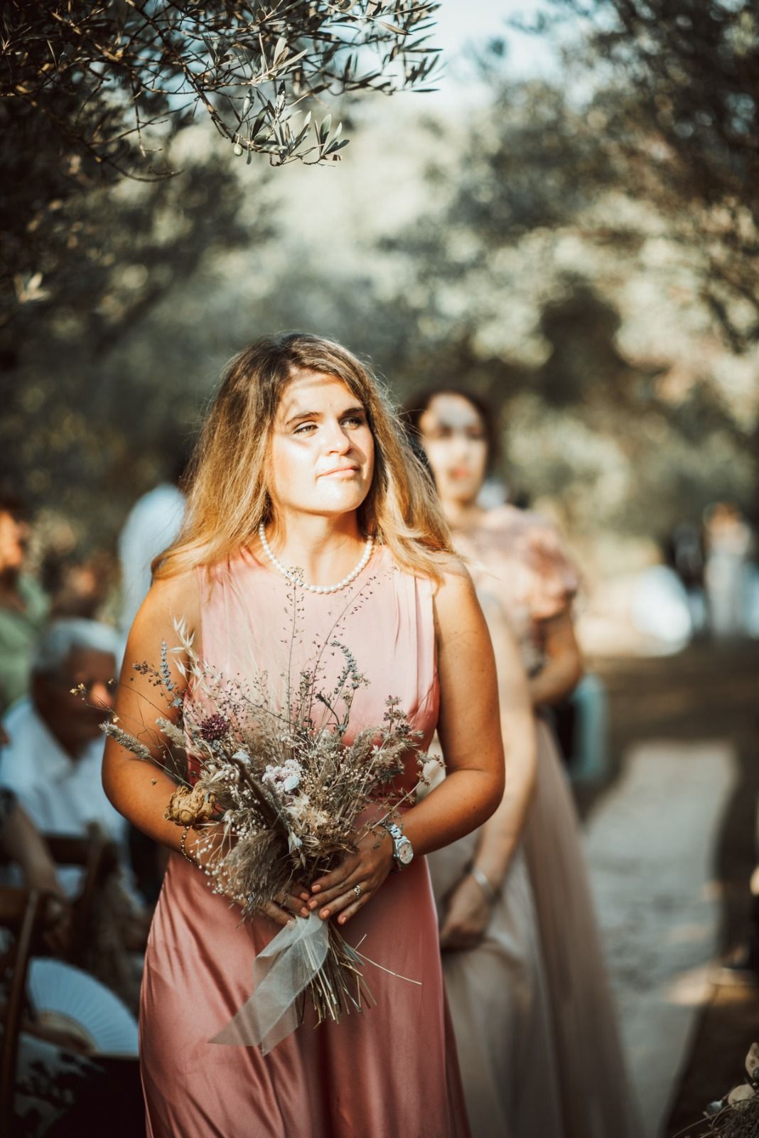 Una donna con un abito rosa porta dei fiori e cammina all'aperto; un'altra donna con un abito la segue.