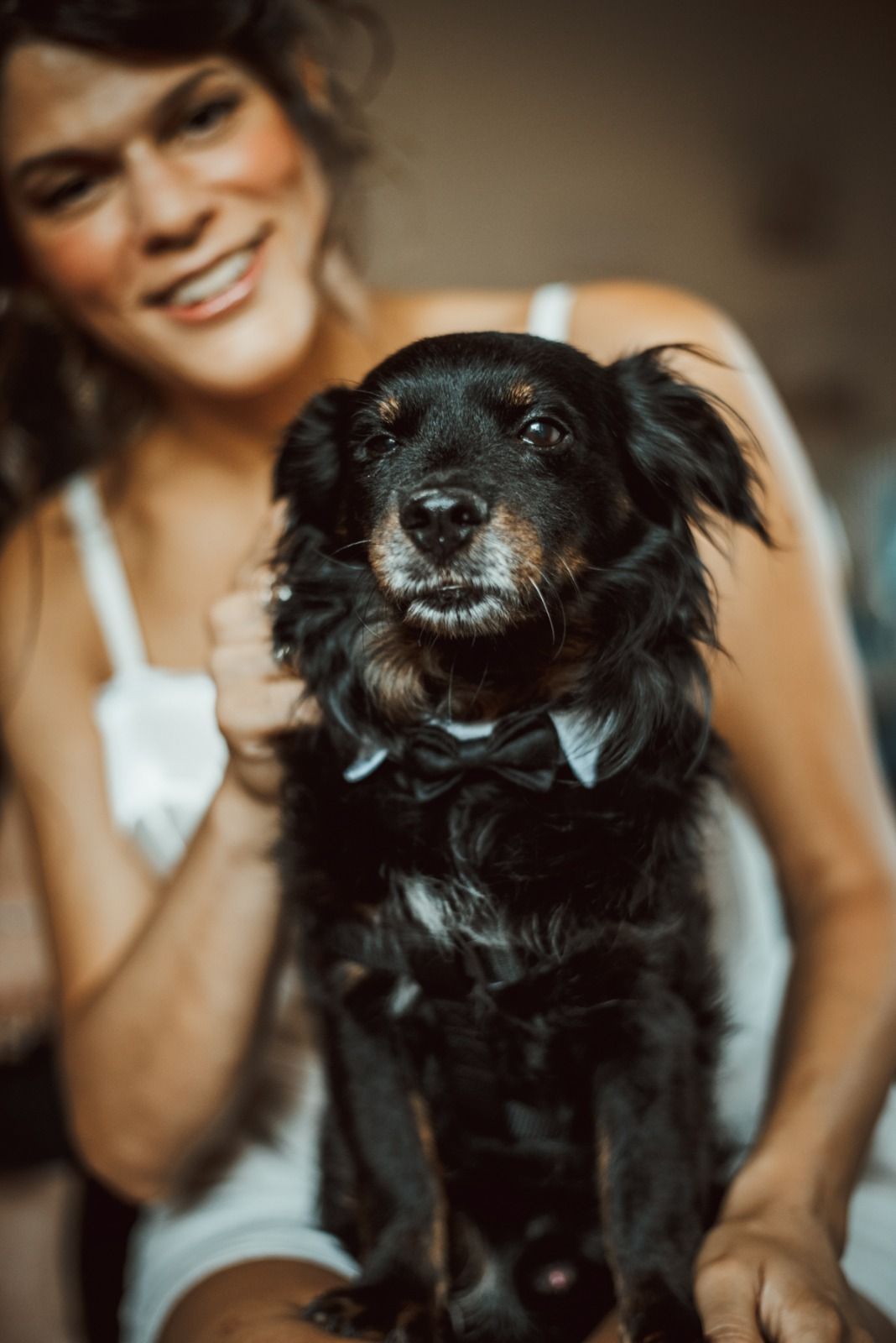 Un cane nero con un papillon è seduto accanto a una donna, entrambi sorridenti. All'interno, luci calde.