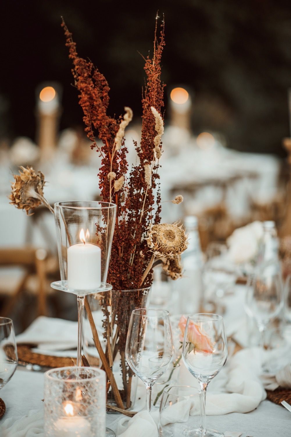 Centrotavola con fiori secchi, candele e bicchieri su un tavolo apparecchiato per un evento. Toni della terra e luci calde.