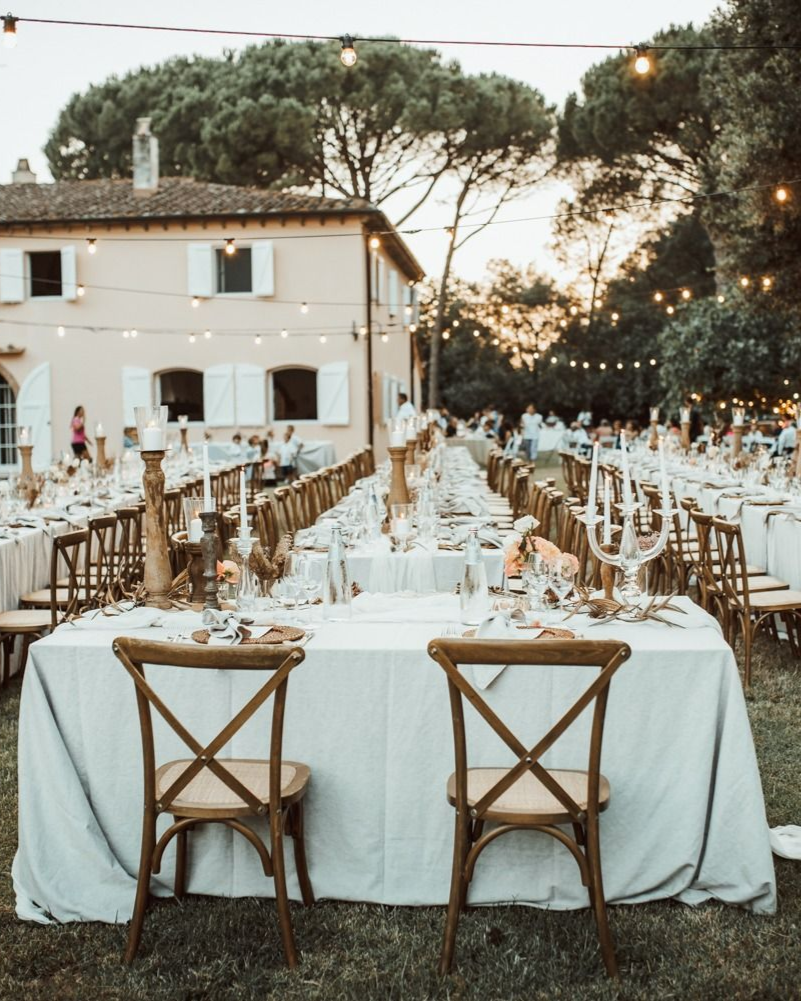 Ricevimento di nozze all'aperto, lunghi tavoli con tovaglie bianche, sedie in legno, luci a filo e una villa toscana.