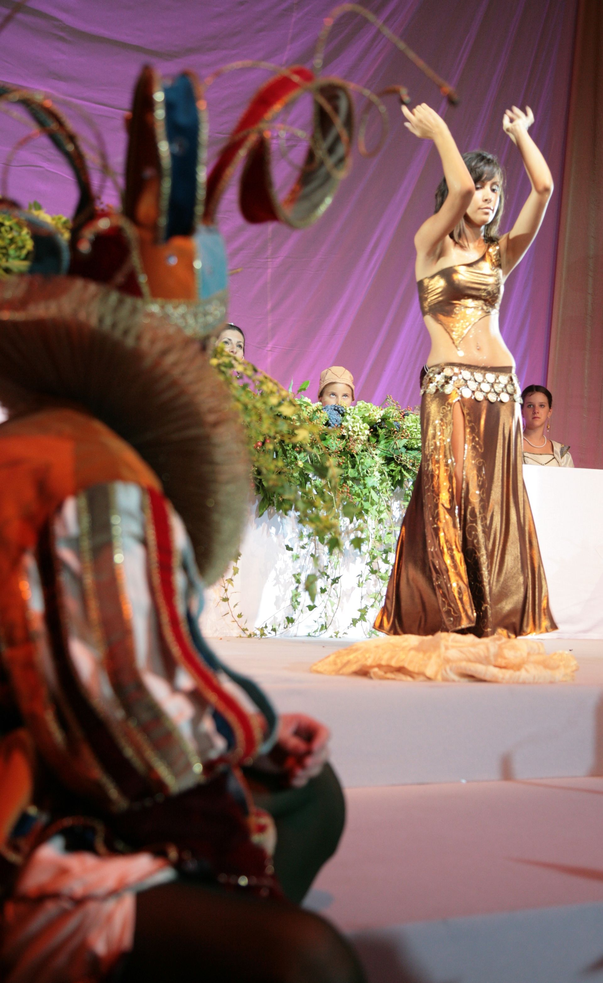 Una danzatrice del ventre in abito dorato si esibisce sul palco, con le braccia alzate. Sullo sfondo: viola e verde. Una figura in costume è in primo piano.