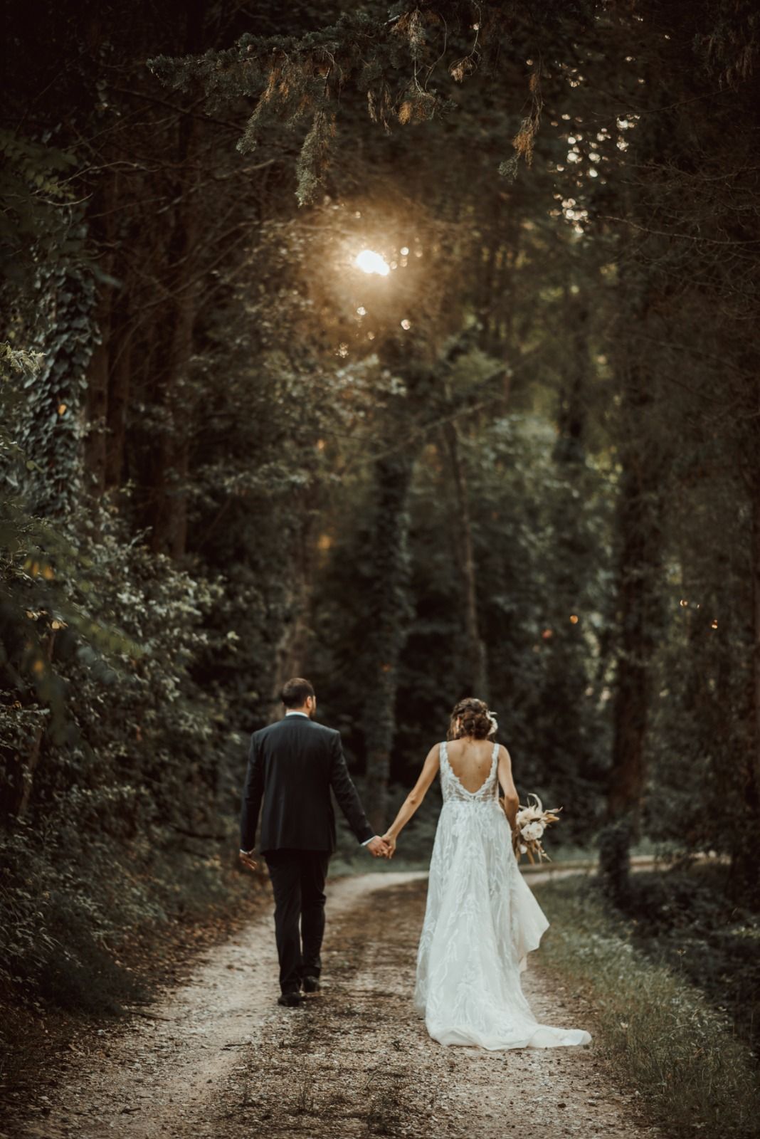 Una coppia in abito da sposa cammina mano nella mano lungo un sentiero nel bosco verso la luce.