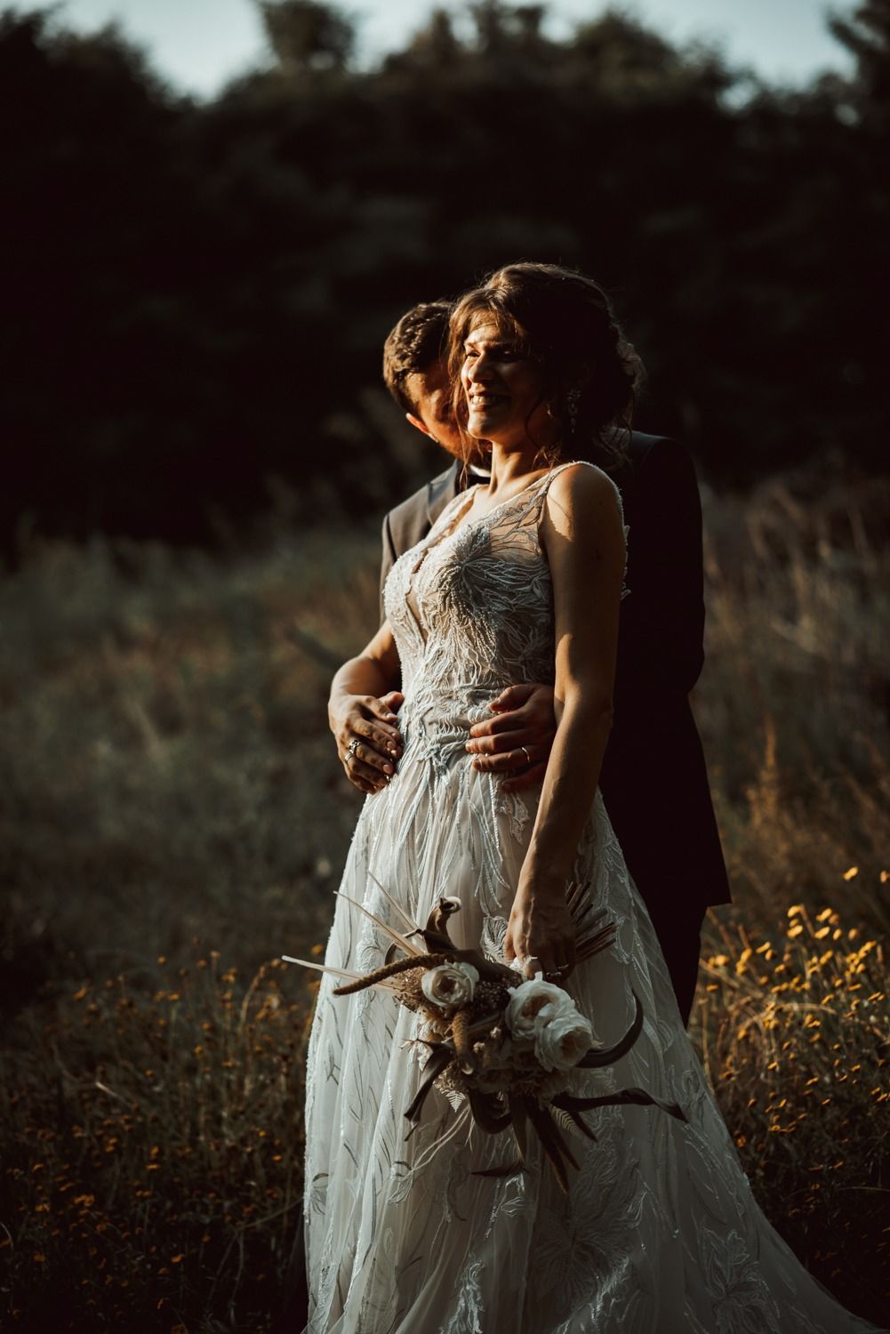 Coppia che si abbraccia in un campo; la sposa in abito bianco, lo sposo in completo. Luce del sole, fiori.