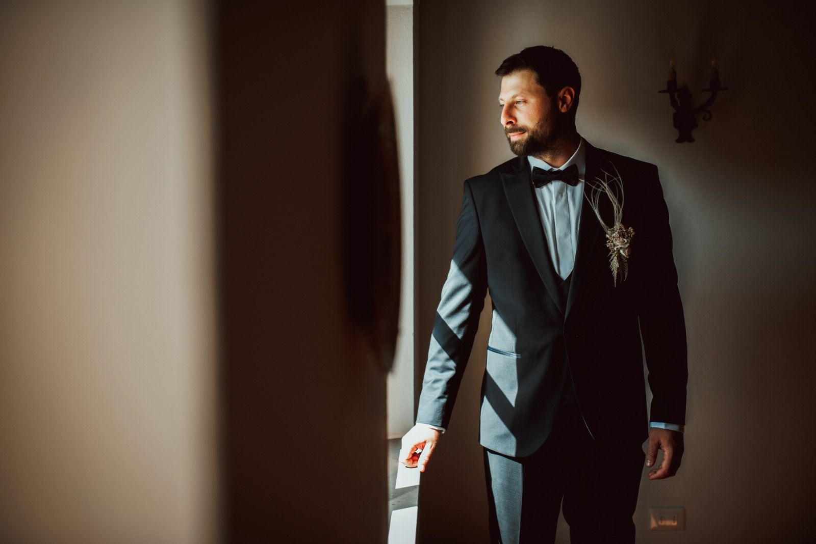 Uomo in abito nero e papillon, in piedi alla luce del sole, che guarda altrove.
