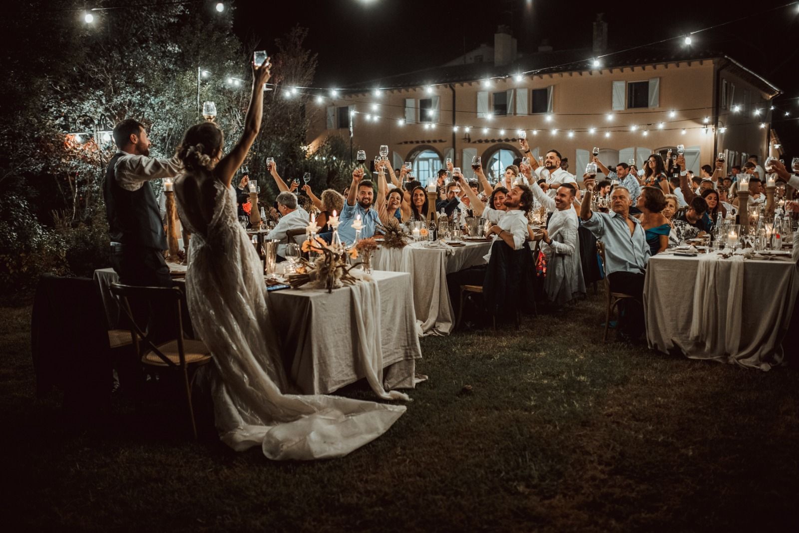 Ricevimento di nozze di sera; la sposa alza lo champagne, la folla applaude, i tavoli sono apparecchiati all'aperto con luci.