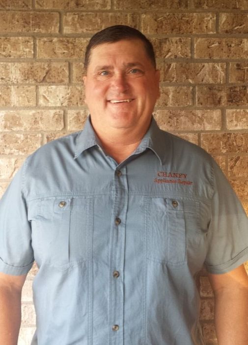 Man standing against a brick wall, wearing a light blue button-up shirt and smiling.