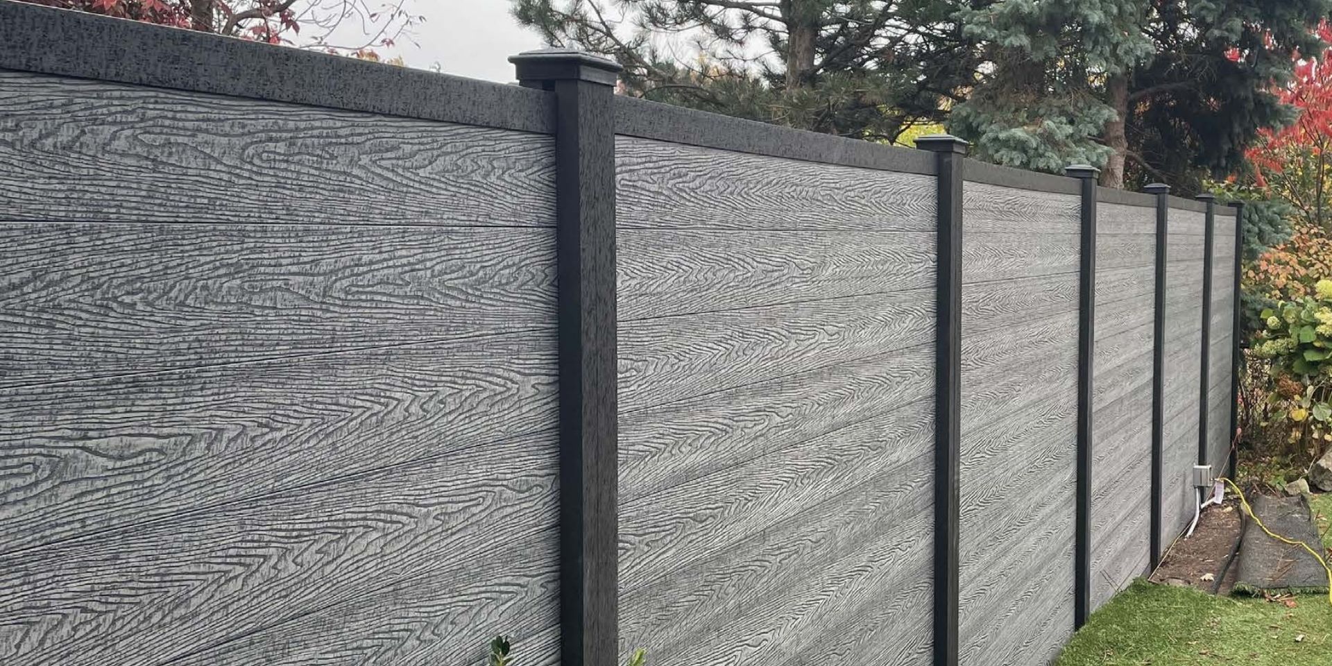 Gray privacy fence with black posts along a yard, backed by trees and foliage