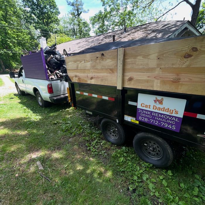 White pickup towing a black trailer loaded with lumber in a grassy yard under trees.