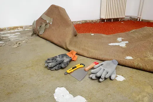 Damaged carpet with peeling floor corner, trowel, utility knife, gloves, and debris on the floor.