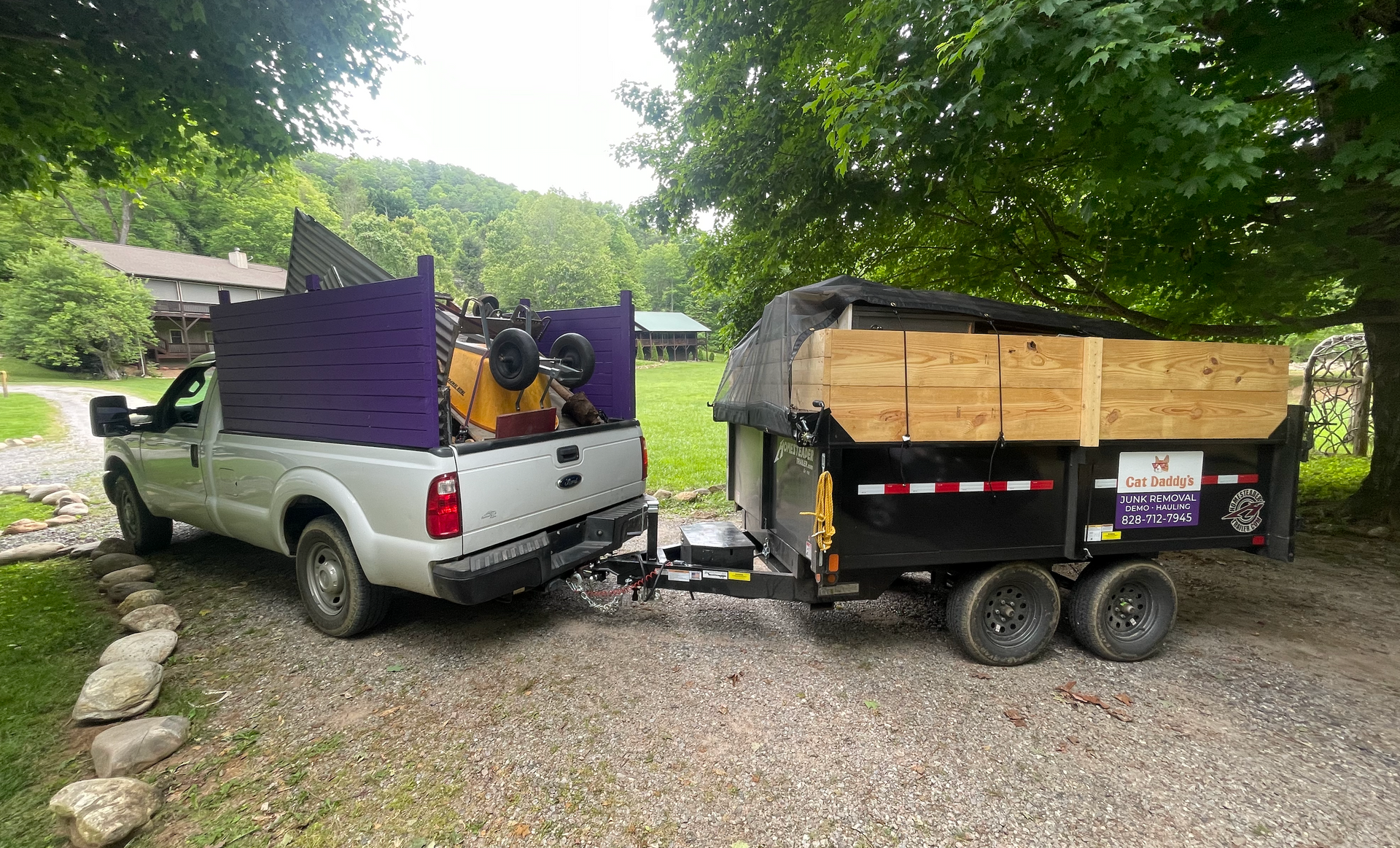 Silver pickup truck hitched to a black dump trailer piled with wood in a gravel driveway.