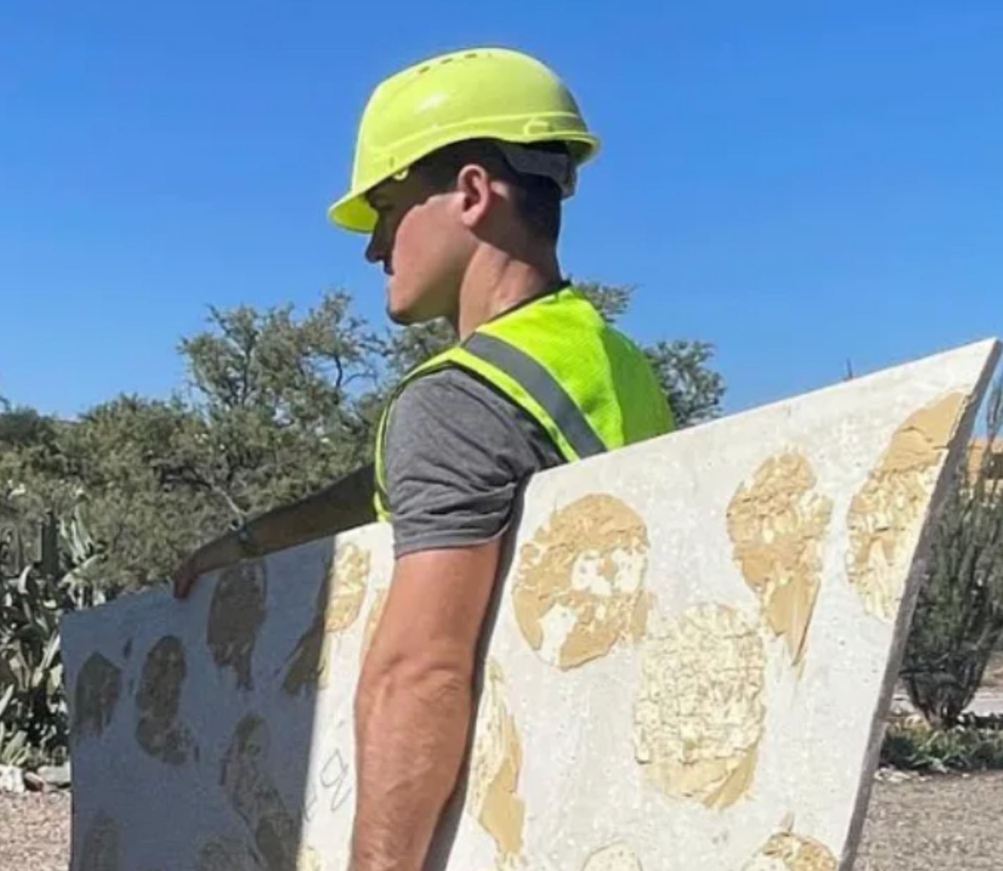 Worker in a neon hard hat and reflective vest carrying large white panels outdoors under a blue sky