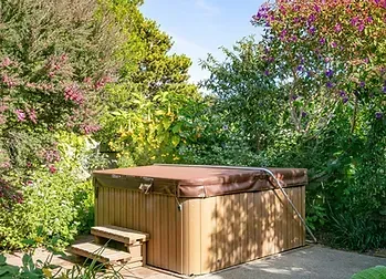 Wooden hot tub on a sunlit patio surrounded by lush green and pink flowering trees.