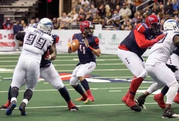 Football player in a blue and red uniform prepares to pass the ball while being pressured by opposing players.