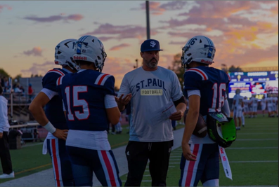 Football coach talking to players on a field. Coach wears a St. Paul Football shirt, players in blue uniforms. Sunset in the background.