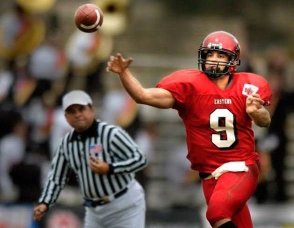 Football player in red uniform throws the ball on a field; referee watches.