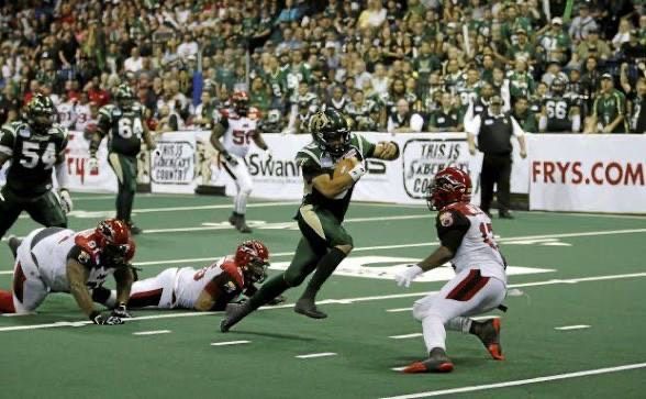Football player in green running towards end zone, pursued by red team. Crowd in background.