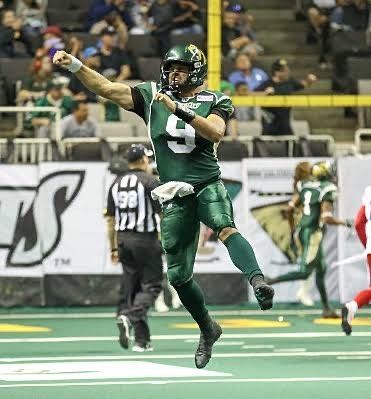 Football player in green uniform celebrates a touchdown, jumping with arm raised. Indoor stadium setting.