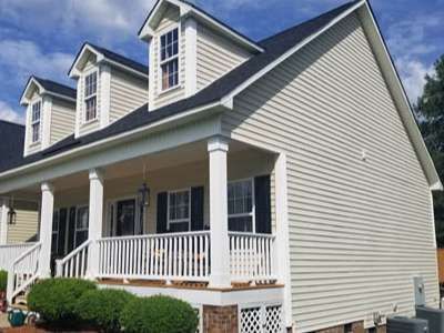 Tan house with black roof, white porch and columns, three dormer windows, green bushes.