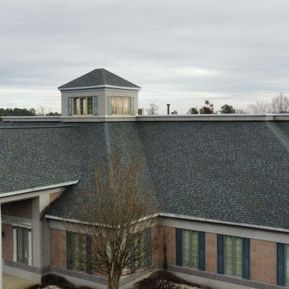 Building with a dark gray shingle roof and a small tower, overcast sky.