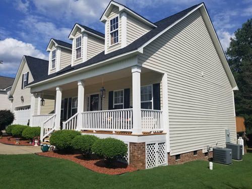 Two-story house with porch, dormers, and black roof. Beige siding, green lawn, and shrubbery.