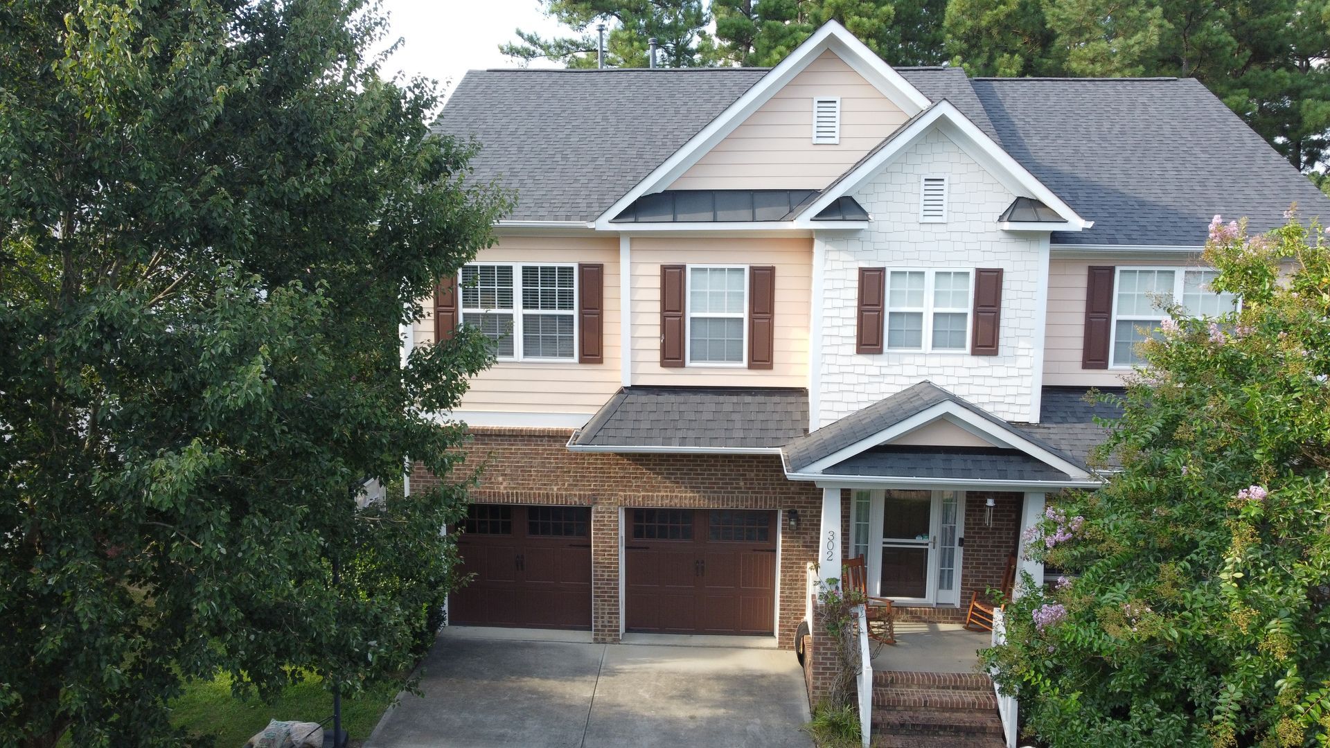 Two-story house with brick accents, brown garage doors, and a small porch; surrounded by green trees.