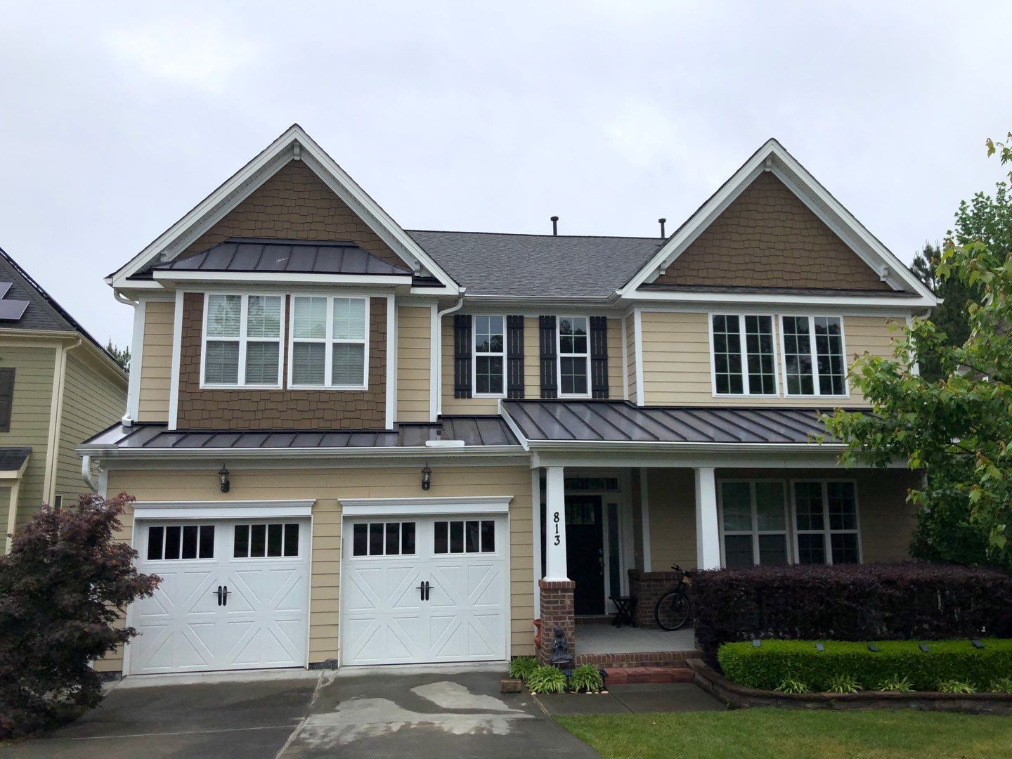 Two-story house with beige siding, brown accents, white trim, and a black roof on a cloudy day.