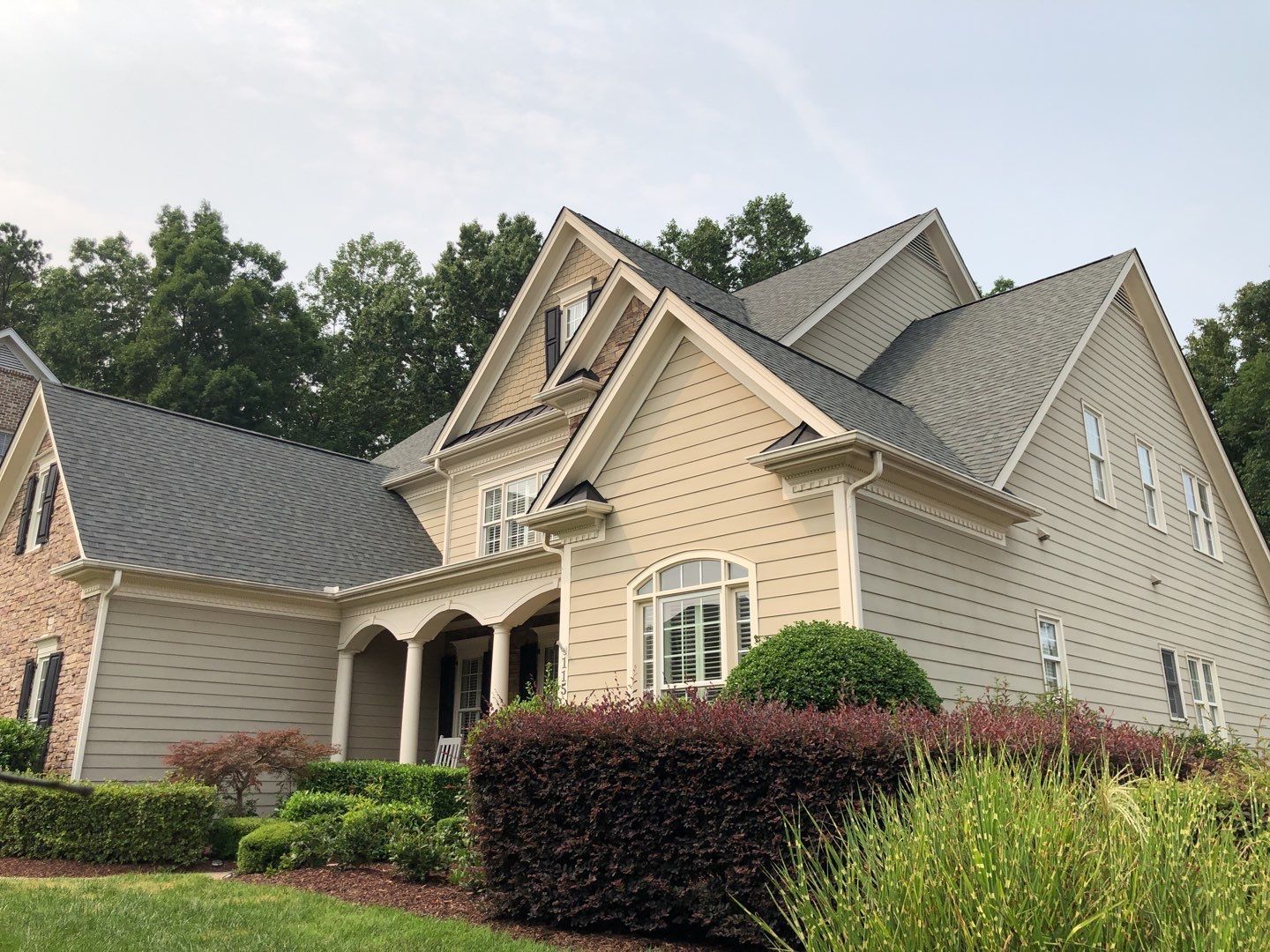 Beige and brick house with gray roof, black shutters, and well-maintained landscaping.