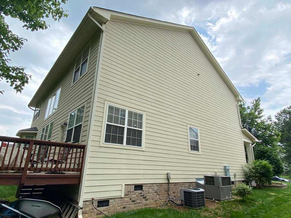 Beige two-story house with a wooden deck, windows, and air conditioning units on the side, under a cloudy sky.