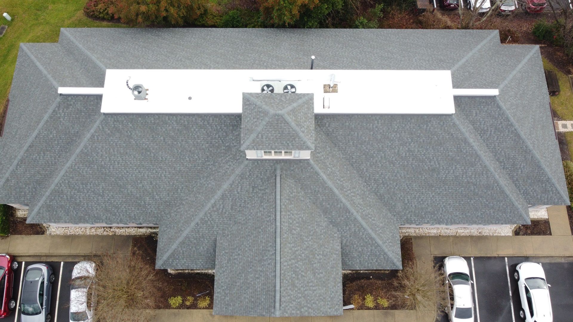 Aerial view of a building with a gray asphalt shingle roof and a white flat roof section. Cars are parked nearby.