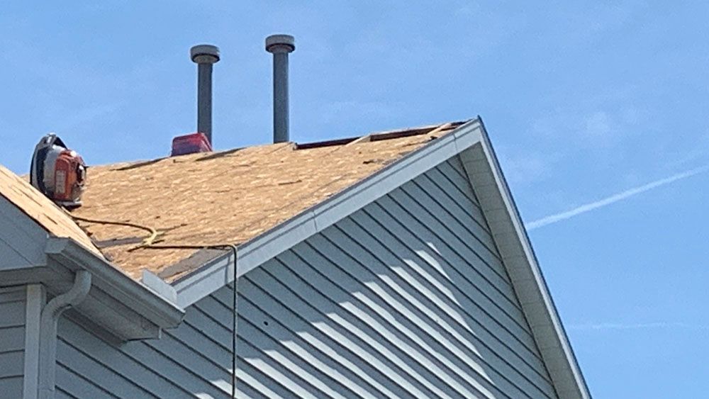 Partially shingled roof with two chimneys and blue siding. A power tool sits on the roof.