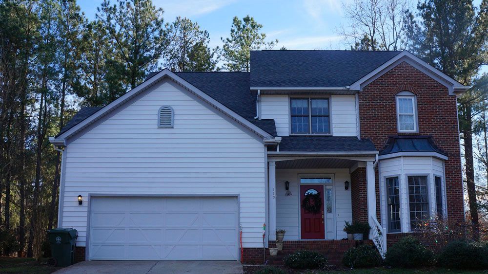 Two-story house with white siding and brick facade, red door, garage, and trees in background.