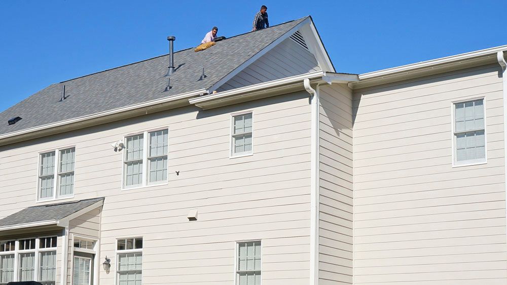 Two people on a gray shingled roof of a beige two-story house, clear blue sky.
