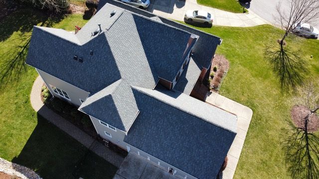 Overhead view of a gray-roofed house with green lawn, driveway, and surrounding trees.
