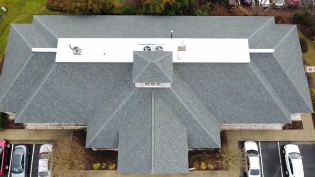 Overhead view of a building with a gray shingle roof and white flat roof section, parking lot below.