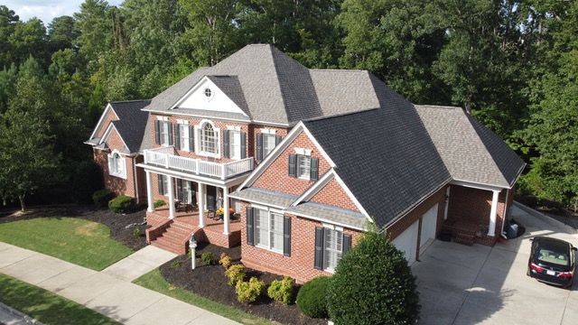 Two-story brick house with a gray roof and white trim, set among trees and a paved driveway.