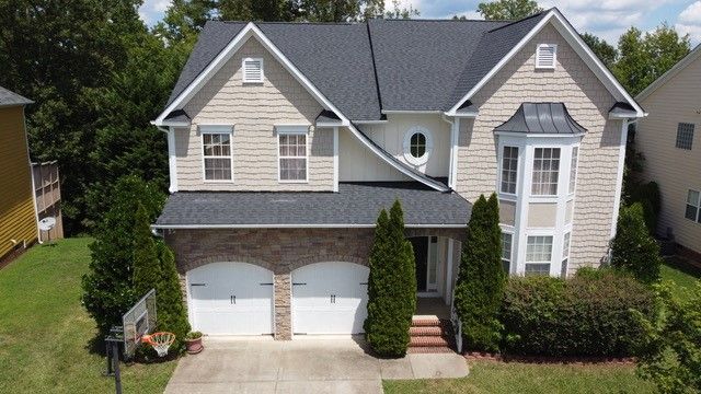 Two-story house with tan siding, brick facade, white garage doors, and a dark gray roof.