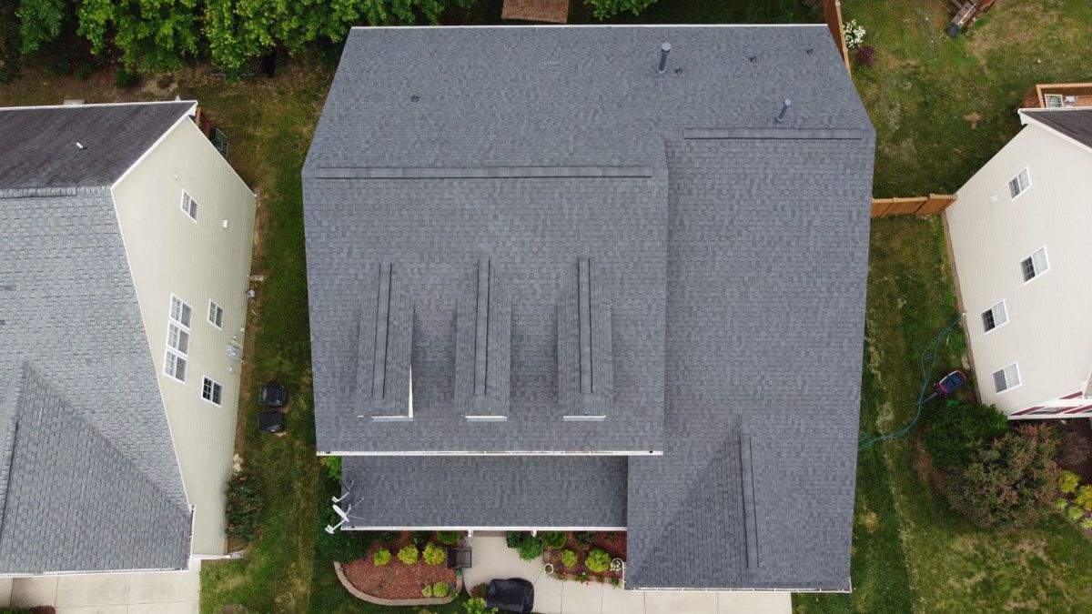 Overhead view of a house with a gray roof, chimneys, and surrounding lawns, trees, and other houses.