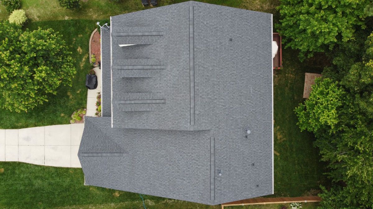 Overhead view of a house with a gray shingled roof, surrounded by green trees and a white concrete walkway.