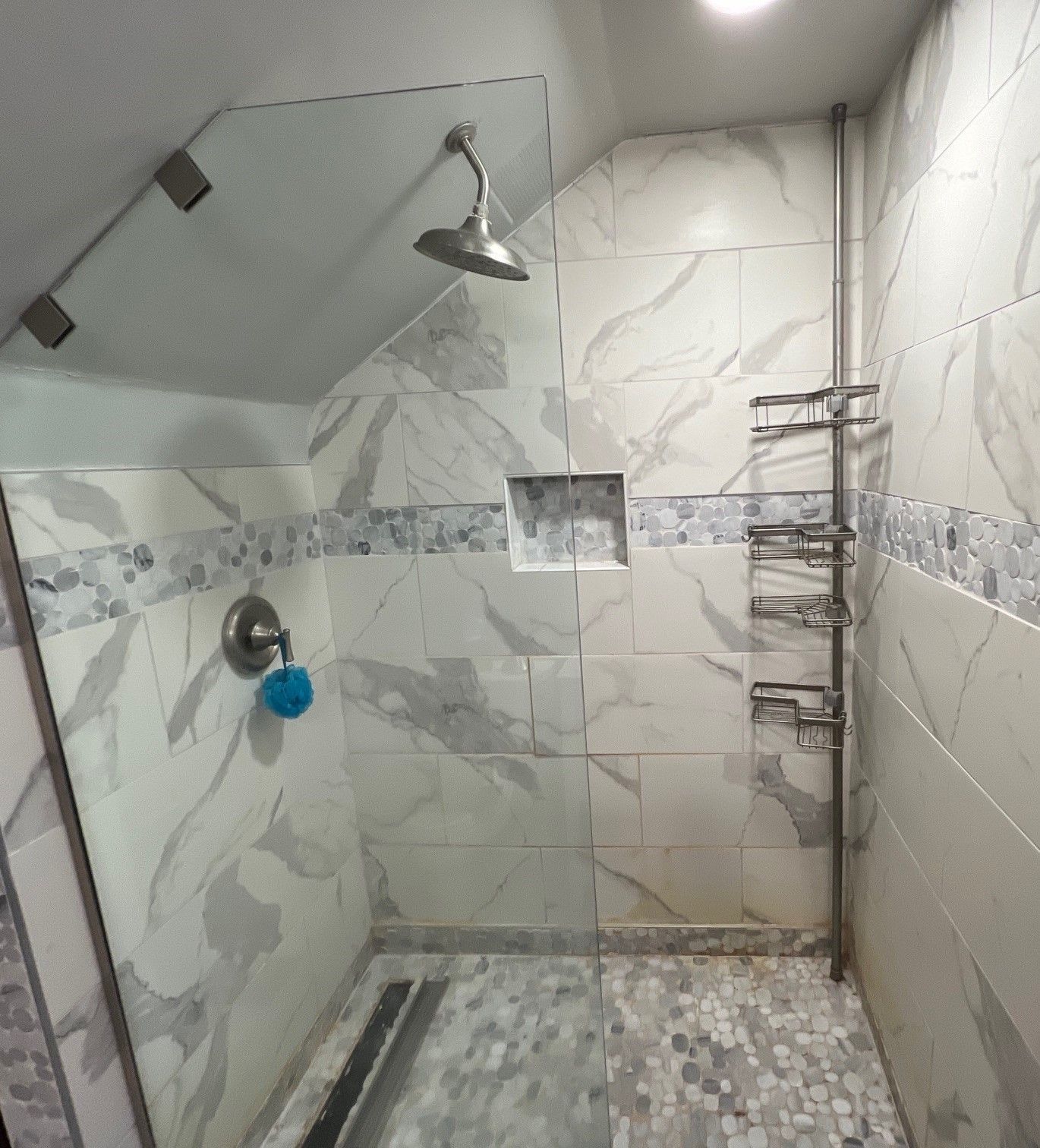 Shower stall with marble-look tiles, glass door, rain shower head, shelf, and mosaic floor.