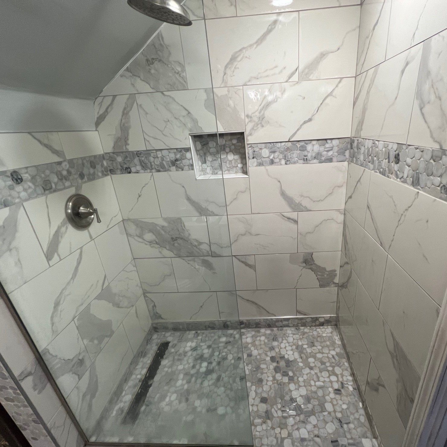 Tiled shower with gray marble-look walls, a pebble floor, and built-in shelf.