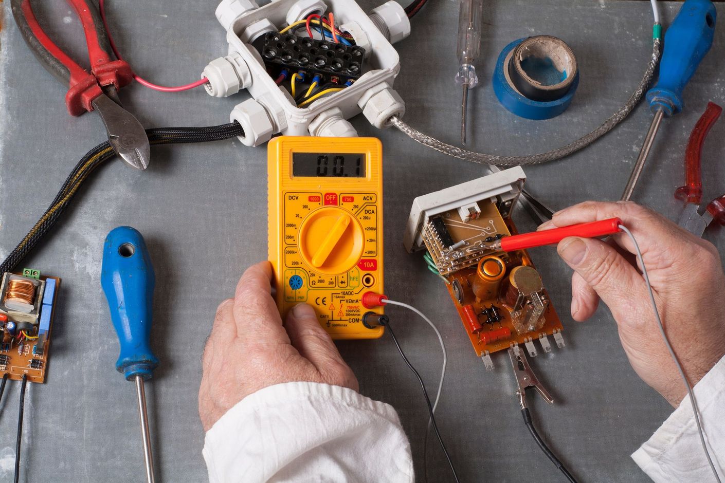 Electrician using digital multimeter to test circuit board with electrical wiring tools.