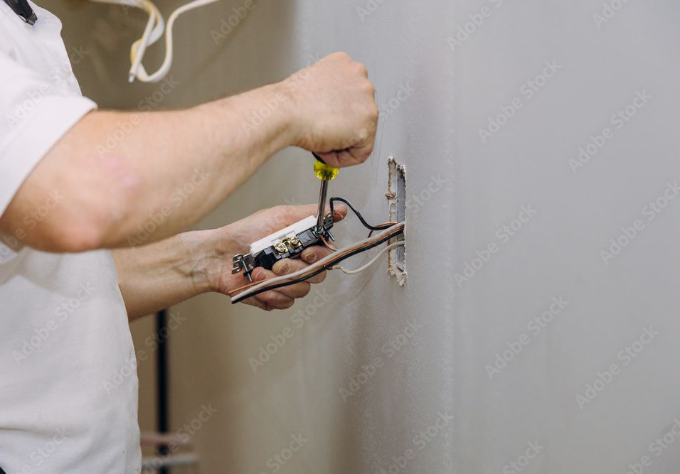 Hand testing an electrical outlet with red and black probes. Wires are visible, wall is white.