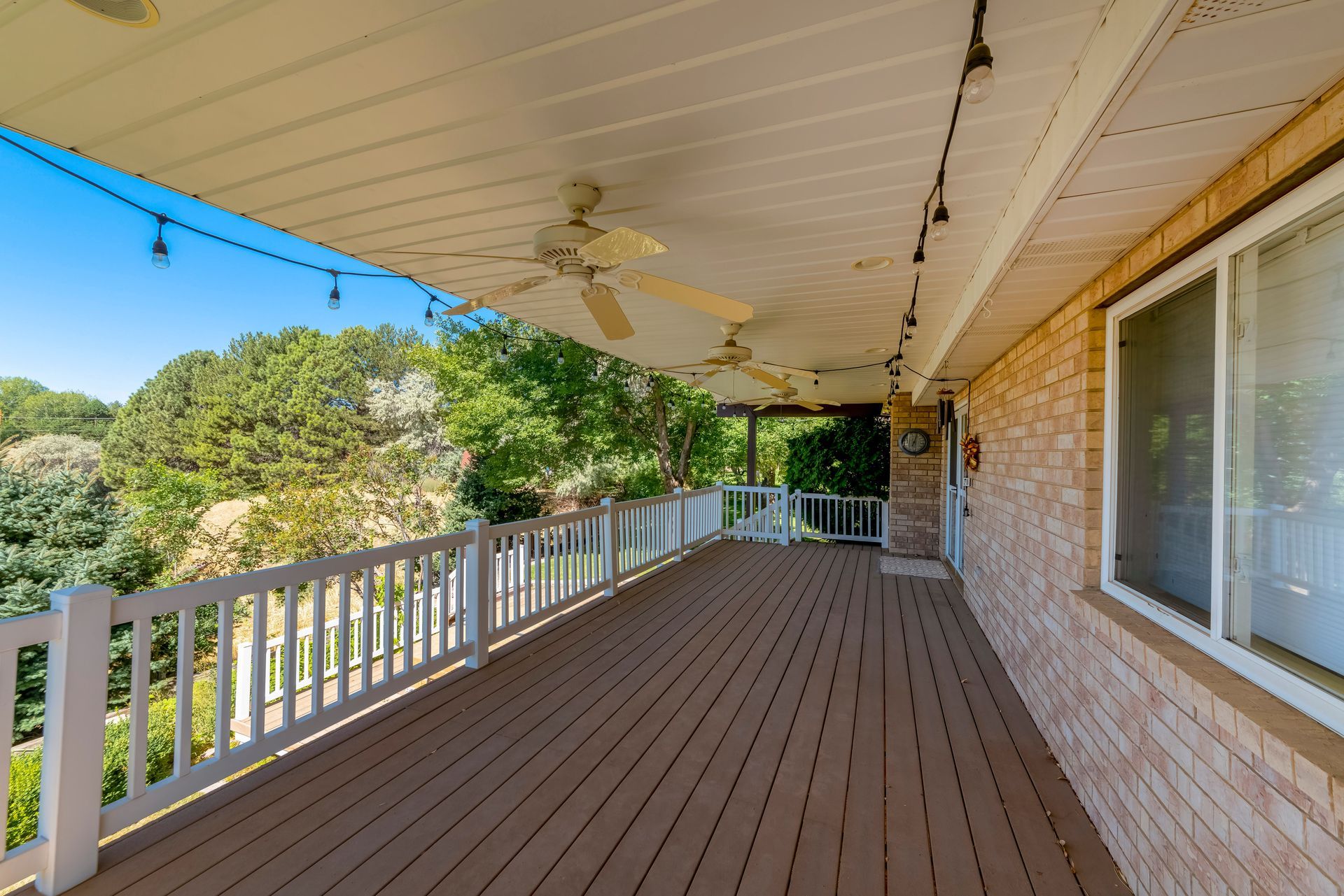 Covered wooden deck with white railing, string lights, and ceiling fans. A window and brick wall are on the right.