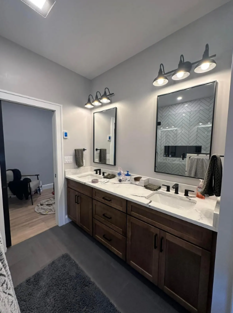 Hand inserting a white outlet cover into an electrical outlet. Beige wall. Bathroom with double vanity, dark wood cabinets, light gray walls, rectangular mirrors, and black fixtures.