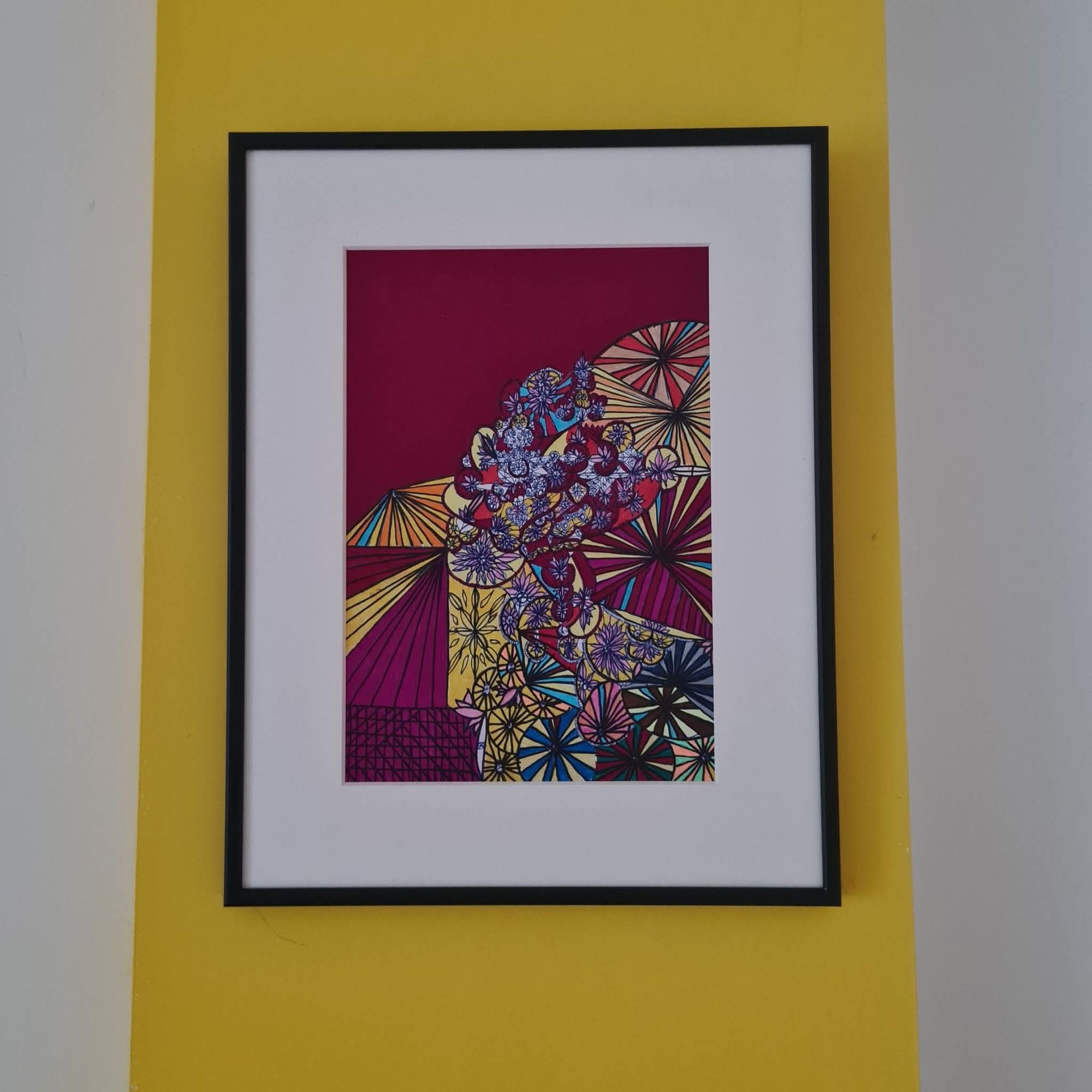 Ingelijst kunstwerk met kleurrijk bloemmotief op een donkerrode achtergrond, gemonteerd op een witte passe-partout, opgehangen tegen een gele muur.