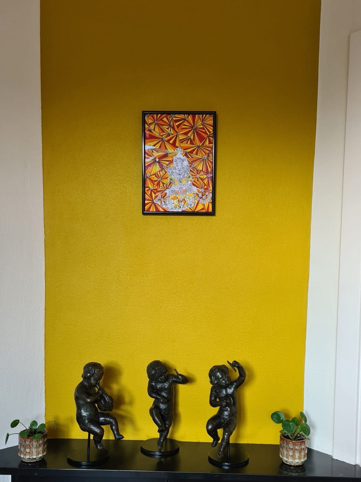 Drie bronzen cherubijnbeelden op een plank onder een ingelijst abstract kunstwerk tegen een gele muur.